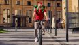 Delivering food to customers. Full length of a young cheerful caucasian male courier with thermo bag riding bicycle along sunny street, for web banner or landing page. Delivery service concept